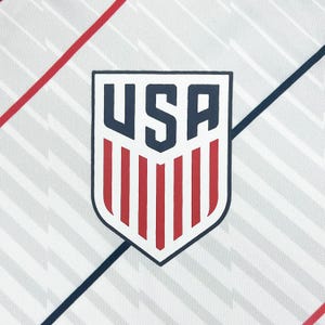 May include: A white fabric with a repeating gray pattern and red and blue diagonal stripes. The image features a shield-shaped emblem with the word "USA" in navy blue, and red vertical stripes at the bottom.
