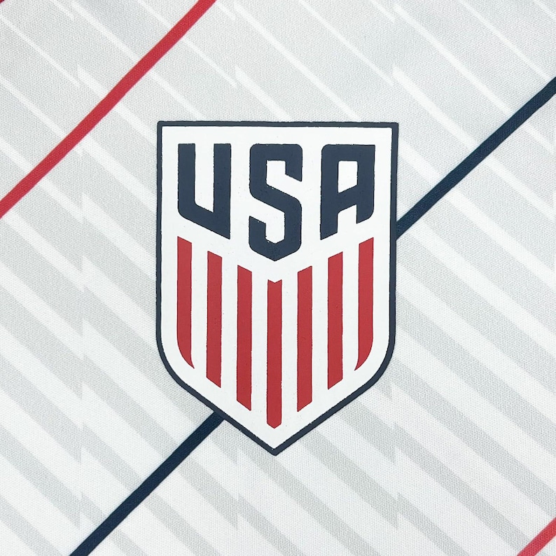 May include: A white fabric with a repeating gray pattern and red and blue diagonal stripes. The image features a shield-shaped emblem with the word "USA" in navy blue, and red vertical stripes at the bottom.