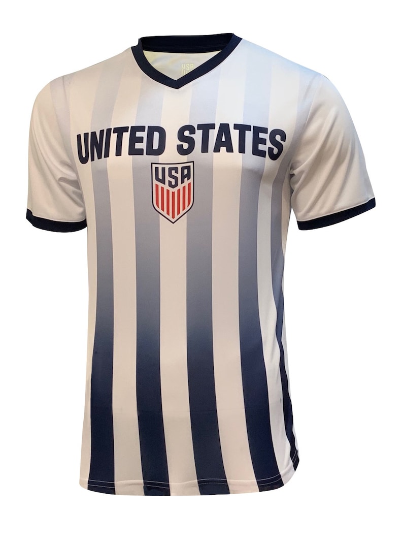 May include: White soccer jersey with blue vertical stripes and a navy blue collar. The jersey has the words "UNITED STATES" printed on the front, with a red, white, and blue shield logo that says "USA" underneath.