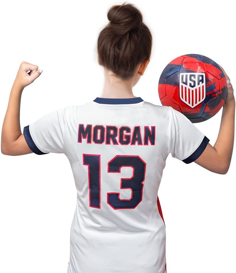 May include: A white soccer jersey with blue trim and the name "MORGAN" and the number "13" on the back. The jersey is being held by a person holding a red, white, and blue soccer ball with the word "USA" on it.