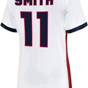 May include: White soccer jersey with a navy blue collar and sleeves. The back of the jersey has the name "SMITH" and the number "11" in navy blue with a red outline.