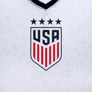 Pode incluir: Uma camisa de futebol branca com um logotipo de escudo vermelho, branco e azul. O escudo tem a palavra "USA" em letras azuis e cinco estrelas brancas acima.