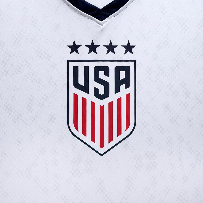 Pode incluir: Uma camisa de futebol branca com um logotipo de escudo vermelho, branco e azul. O escudo tem a palavra "USA" em letras azuis e cinco estrelas brancas acima.