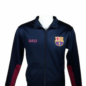 May include: A navy blue zippered jacket with maroon accents on the sleeves. The jacket has the word "BARCA" on the left side and the FC Barcelona logo on the right side.