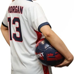 Puede incluir: Una camiseta de f&uacute;tbol blanca con ribetes rojos y azules. La camiseta tiene el nombre "MORGAN" y el n&uacute;mero "13" en la espalda. La persona est&aacute; sosteniendo un bal&oacute;n de f&uacute;tbol con un dise&ntilde;o negro, rojo y blanco.