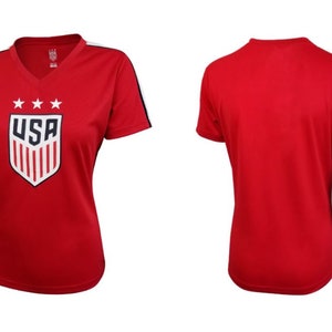 Pode incluir: Camiseta vermelha de manga curta com um bras&atilde;o dos EUA branco e azul com estrelas e listras. A camiseta tem listras brancas nas mangas.