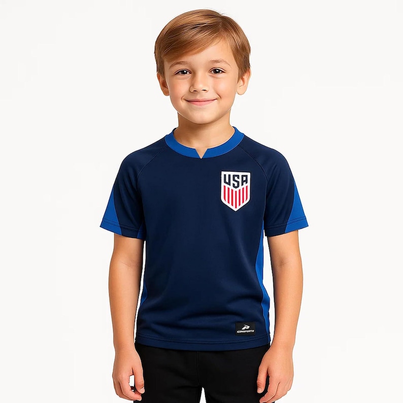 May include: A navy blue youth soccer jersey with blue accents on the sleeves and collar. The jersey features the USA logo on the chest. The boy is wearing black pants.