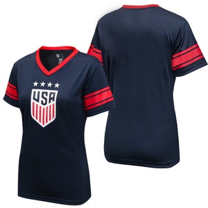 May include: Navy blue short-sleeved t-shirt with red and white stripes on the sleeves. The front of the shirt features a white shield with red and white stripes and the word "USA" in white letters. There are five white stars above the shield.