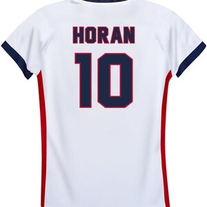 Pode incluir: Camisola de futebol branca com guarni&ccedil;&otilde;es vermelhas e azuis. A parte de tr&aacute;s da camisola tem o nome "HORAN" e o n&uacute;mero "10" impresso em azul com um contorno vermelho.