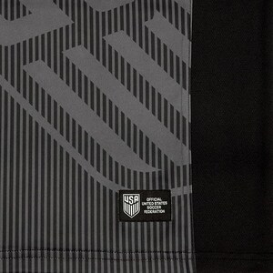Puede incluir: Una camiseta de f&uacute;tbol negra y gris con un patr&oacute;n geom&eacute;trico. La camiseta tiene un parche en la parte delantera que dice "Official United States Soccer Federation".