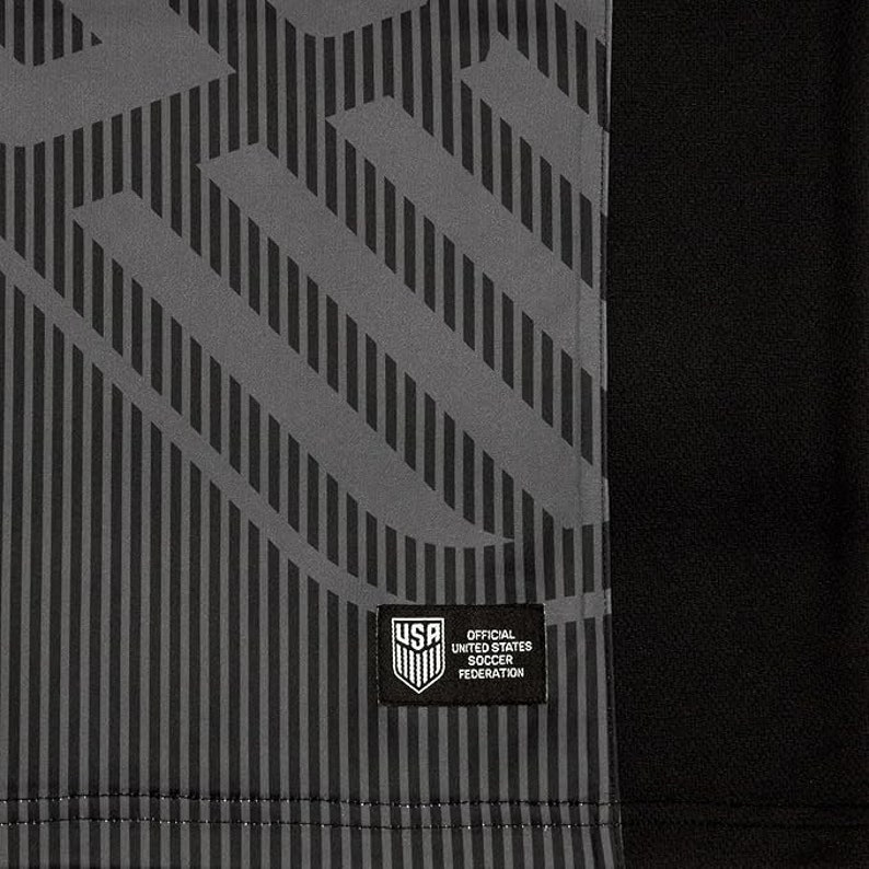 Puede incluir: Una camiseta de f&uacute;tbol negra y gris con un patr&oacute;n geom&eacute;trico. La camiseta tiene un parche en la parte delantera que dice "Official United States Soccer Federation".