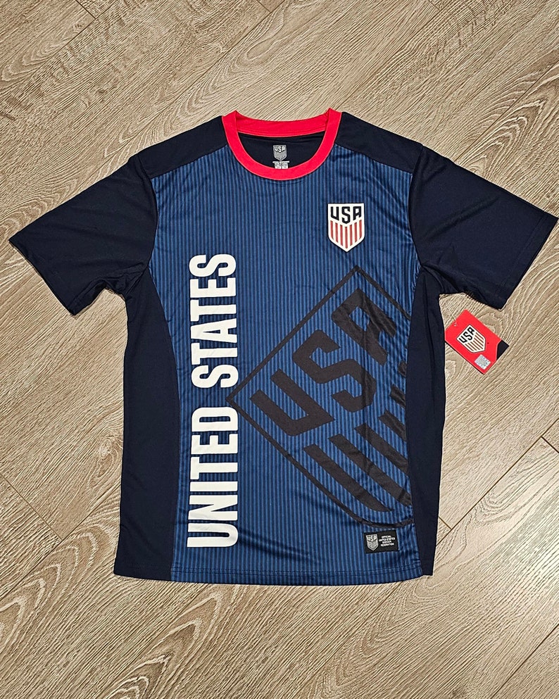 May include: A navy blue soccer jersey with a white "UNITED STATES" and "USA" graphic on the front. The jersey has a red and white trim around the neck and a red "USA" logo on the chest.