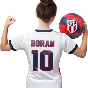 Pode incluir: Camisola de futebol branca com detalhes azuis e vermelhos. Na parte de tr&aacute;s da camisola, o nome "HORAN" e o n&uacute;mero "10" est&atilde;o impressos em azul com contorno vermelho. Uma pessoa est&aacute; vestindo a camisola e segurando uma bola de futebol vermelha, branca e azul com a palavra "USA" impressa nela.