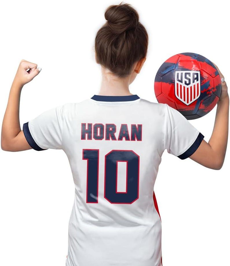 Pode incluir: Camisola de futebol branca com detalhes azuis e vermelhos. Na parte de tr&aacute;s da camisola, o nome "HORAN" e o n&uacute;mero "10" est&atilde;o impressos em azul com contorno vermelho. Uma pessoa est&aacute; vestindo a camisola e segurando uma bola de futebol vermelha, branca e azul com a palavra "USA" impressa nela.