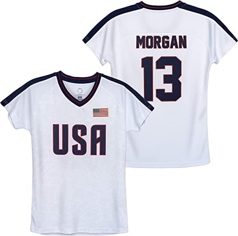 May include: White soccer jersey with blue and red accents. The jersey has the word "USA" on the front and a small American flag. The back of the jersey has the name "MORGAN" and the number "13" in blue.