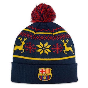 May include: A navy blue knitted beanie with a yellow and red patterned design featuring snowflakes and reindeer. The beanie has a red pom pom on top and a navy blue patch with the FC Barcelona logo on the front.