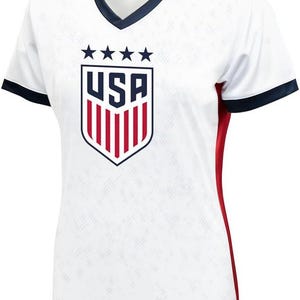 K&ouml;nnte beinhalten: Wei&szlig;es Fu&szlig;balltrikot mit einem blauen und roten USA-Wappen und f&uuml;nf Sternen. Das Trikot hat einen marineblauen Kragen und einen Saum an den &Auml;rmeln. Die Seiten des Trikots haben rote Akzente.