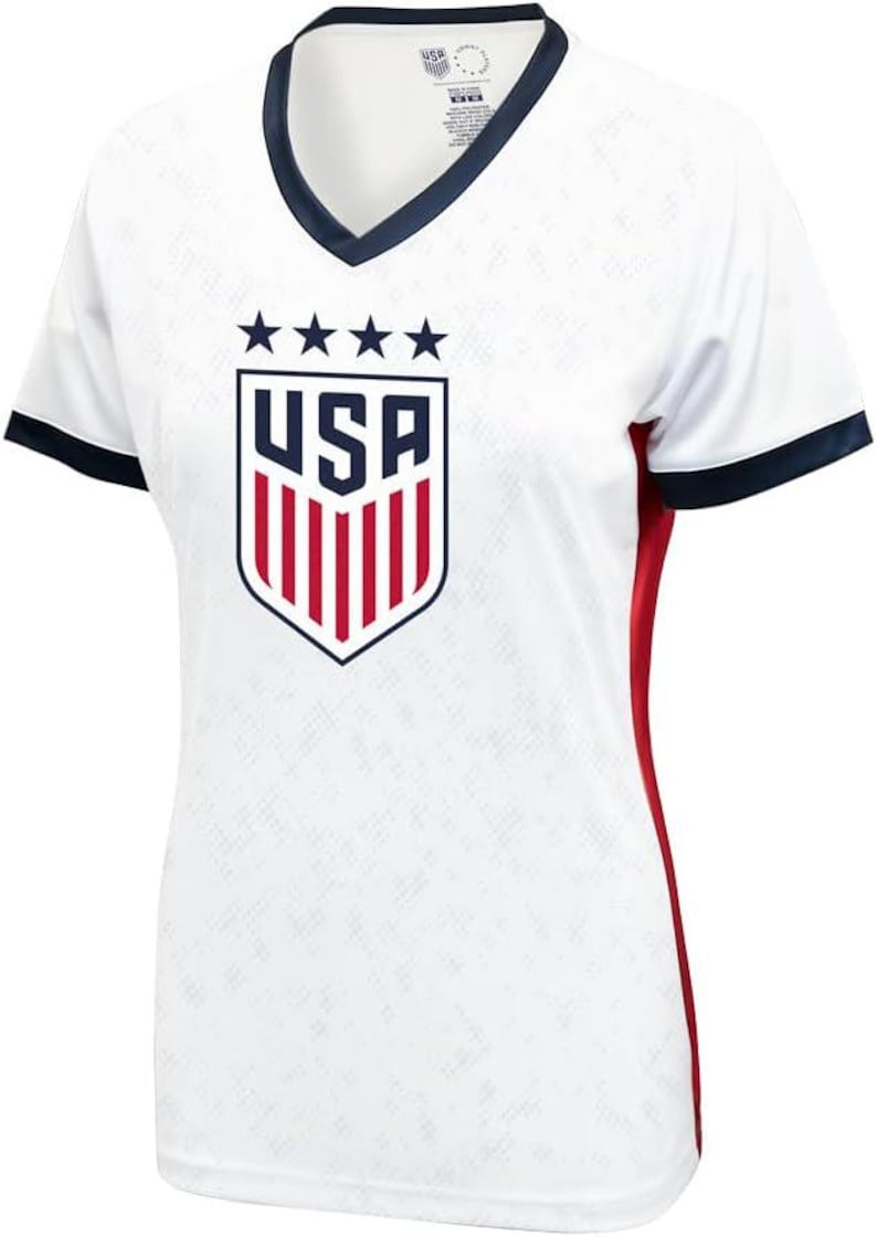 K&ouml;nnte beinhalten: Wei&szlig;es Fu&szlig;balltrikot mit einem blauen und roten USA-Wappen und f&uuml;nf Sternen. Das Trikot hat einen marineblauen Kragen und einen Saum an den &Auml;rmeln. Die Seiten des Trikots haben rote Akzente.
