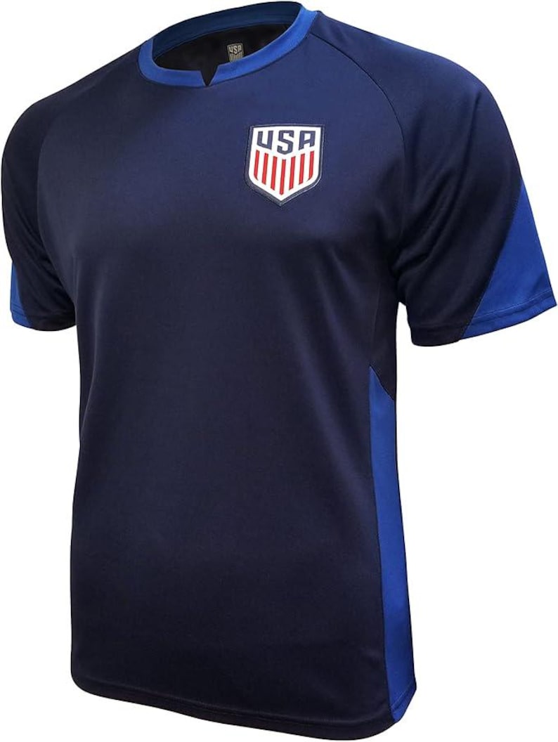 Puede incluir: Una camiseta de f&uacute;tbol azul marino con un logotipo de EE. UU. azul y blanco en la parte delantera. La camiseta tiene detalles azules en las mangas y los lados.