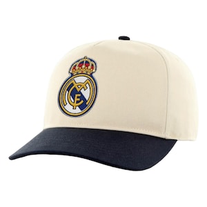 May include: Cream-colored baseball cap with a navy blue brim. The cap features the Real Madrid logo embroidered on the front. The logo includes a crown, a circular emblem with the letters "RM", and a gold border.