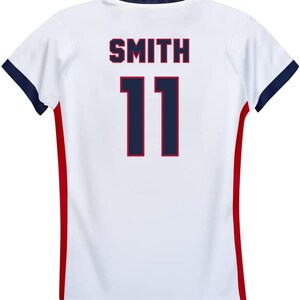May include: White athletic jersey with the name "SMITH" and the number "11" in navy blue. The jersey has navy blue trim on the sleeves and a red stripe down the sides.