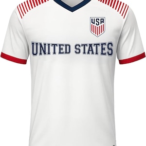May include: White soccer jersey with red and blue accents. The jersey features a V-neck collar, red stripes on the shoulders, and red trim on the sleeves. The words "UNITED STATES" are across the chest, with the "USA" logo on the upper right.