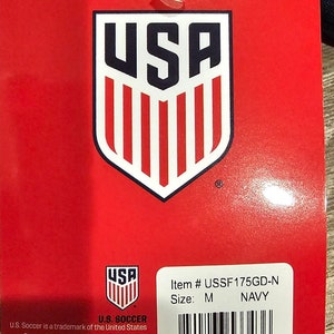 May include: Red tag with a white and red USA logo. The tag has the text "Item # USSF175GD-N Size: M NAVY" and a barcode. The text "U.S. SOCCER" is at the bottom of the tag with copyright information.