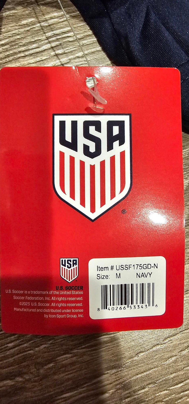May include: Red tag with a white and red USA logo. The tag has the text "Item # USSF175GD-N Size: M NAVY" and a barcode. The text "U.S. SOCCER" is at the bottom of the tag with copyright information.