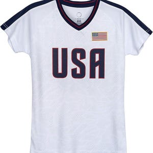 May include: White athletic jersey with a V-neck collar and short sleeves. The jersey features the word "USA" in navy blue block letters, an American flag patch, and navy blue trim.