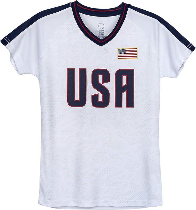 May include: White athletic jersey with a V-neck collar and short sleeves. The jersey features the word "USA" in navy blue block letters, an American flag patch, and navy blue trim.