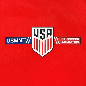 Puede incluir: Primer plano de una tela roja con un emblema blanco, azul y rojo. El emblema presenta la palabra "USA" sobre un escudo con rayas blancas verticales. Debajo, las palabras "USMNT" y "U.S. SOCCER FEDERATION".