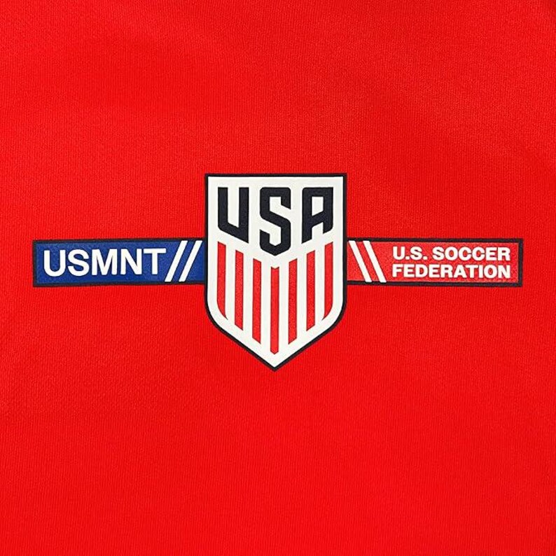 Puede incluir: Primer plano de una tela roja con un emblema blanco, azul y rojo. El emblema presenta la palabra "USA" sobre un escudo con rayas blancas verticales. Debajo, las palabras "USMNT" y "U.S. SOCCER FEDERATION".