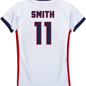 Op de afbeelding: Wit voetbaltenue met rode en donkerblauwe bies. De achterkant van het tenue heeft de naam "SMITH" en het nummer "11" gedrukt in donkerblauw met een rode omtrek.