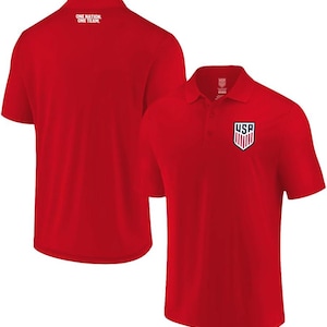 May include: A bright red short-sleeved polo shirt featuring a collar with a button placket. The front displays a USA shield logo in red, white, and blue. The back of the shirt includes the white text "ONE NATION. ONE TEAM." near the neckline.
