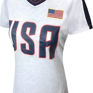 K&ouml;nnte beinhalten: Wei&szlig;es Kurzarm-Trikot mit marineblauem und rotem Besatz. Das Trikot hat ein wei&szlig;-graues geometrisches Muster und das Wort "USA" in Marineblau mit roter Umrandung. Eine kleine amerikanische Flagge befindet sich auf der rechten Brustseite.
