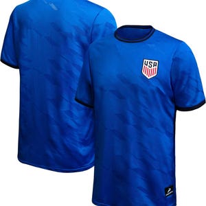 May include: Two blue soccer jerseys. The front jersey has a USA crest on the chest and black trim on the collar and sleeves. The jerseys have a subtle geometric pattern. The second jersey is in the background, showing the back of the shirt.