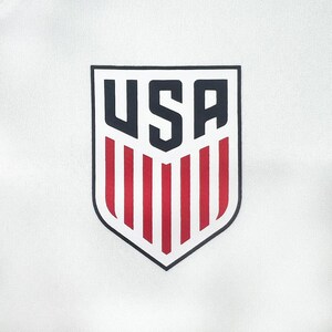May include: White fabric with a shield-shaped emblem. The emblem features the word "USA" in navy blue, above red and white vertical stripes, all outlined in black. The design is centered on the fabric.