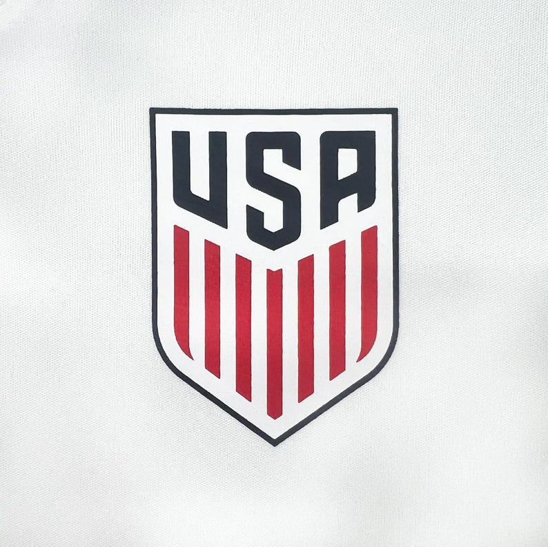 May include: White fabric with a shield-shaped emblem. The emblem features the word "USA" in navy blue, above red and white vertical stripes, all outlined in black. The design is centered on the fabric.