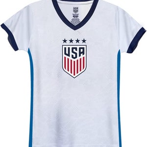 May include: White soccer jersey with blue trim and a navy blue v-neck collar. The jersey features a red, white, and blue shield with the word "USA" in white letters and five stars above the shield.