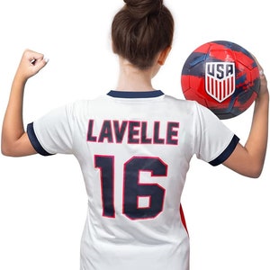 May include: A white soccer jersey with blue trim and the name "Lavelle" and number "16" on the back. The jersey is being held by a person wearing a bun. A red, white, and blue soccer ball with the word "USA" on it is being held in the other hand.
