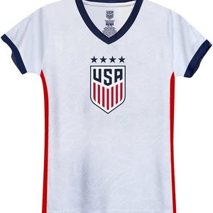 Op de afbeelding: Wit voetbaltenue met rode en blauwe bies. Het tenue heeft een groot USA-wapen met vijf sterren erboven.
