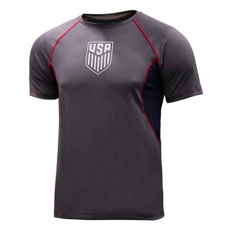 May include: Gray athletic t-shirt with the USA emblem on the chest. The shirt has red trim along the sleeves and shoulders, and a navy blue panel on the side. The fabric has a textured, dotted pattern.