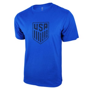 Op de afbeelding: Een koninklijk blauw t-shirt met een zwarte, verweerde afbeelding van het USA-schildlogo.