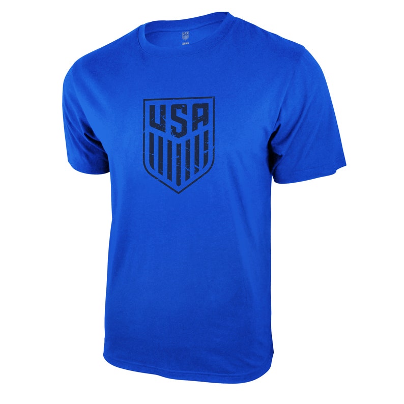 Op de afbeelding: Een koninklijk blauw t-shirt met een zwarte, verweerde afbeelding van het USA-schildlogo.