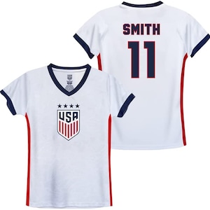 May include: White USA soccer jersey with a V-neck, navy blue trim, and red side panels. The front features the USA crest and stars. The back displays "SMITH" and the number "11" in navy blue.