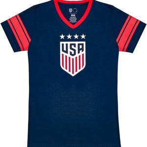 Puede incluir: Una camiseta azul marino con rayas rojas y blancas en las mangas. La camiseta tiene un cuello en V blanco y un ribete de cuello en V rojo. La parte delantera de la camiseta presenta un escudo blanco con la palabra "USA" y cinco estrellas encima.