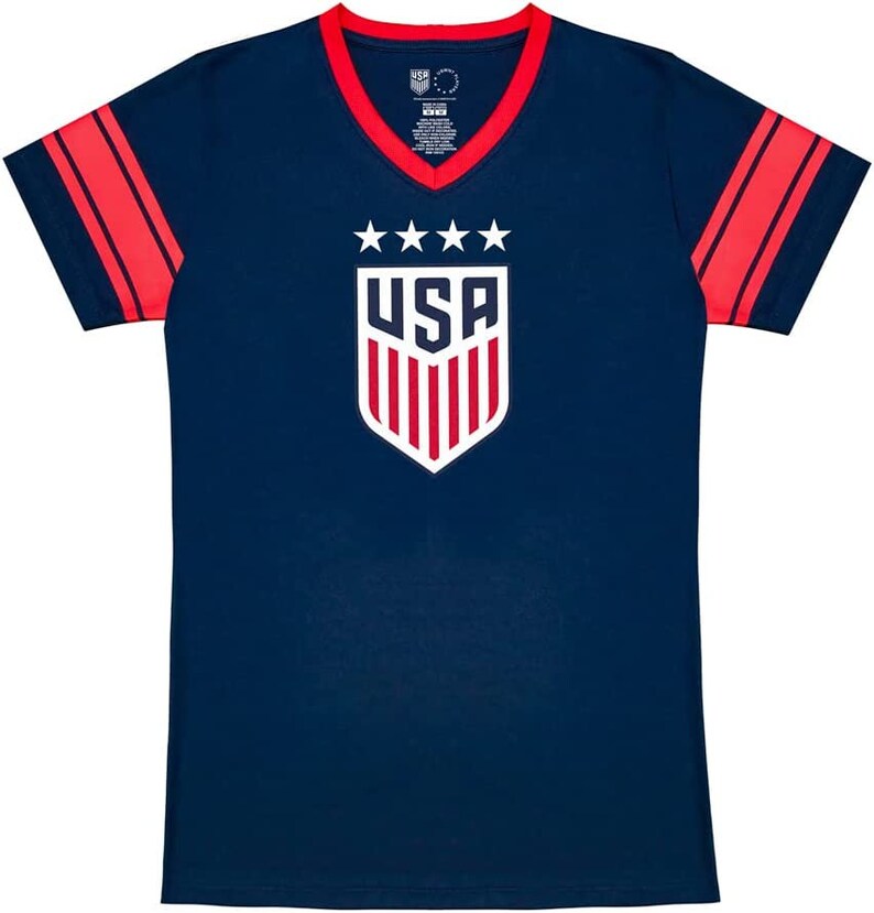 Puede incluir: Una camiseta azul marino con rayas rojas y blancas en las mangas. La camiseta tiene un cuello en V blanco y un ribete de cuello en V rojo. La parte delantera de la camiseta presenta un escudo blanco con la palabra "USA" y cinco estrellas encima.