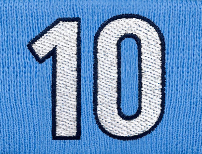 May include: A close-up shot of the number 10 embroidered onto a light blue knitted fabric. The number is white with a navy blue border, displaying a textured, stitched design. The fabric has a knitted appearance.