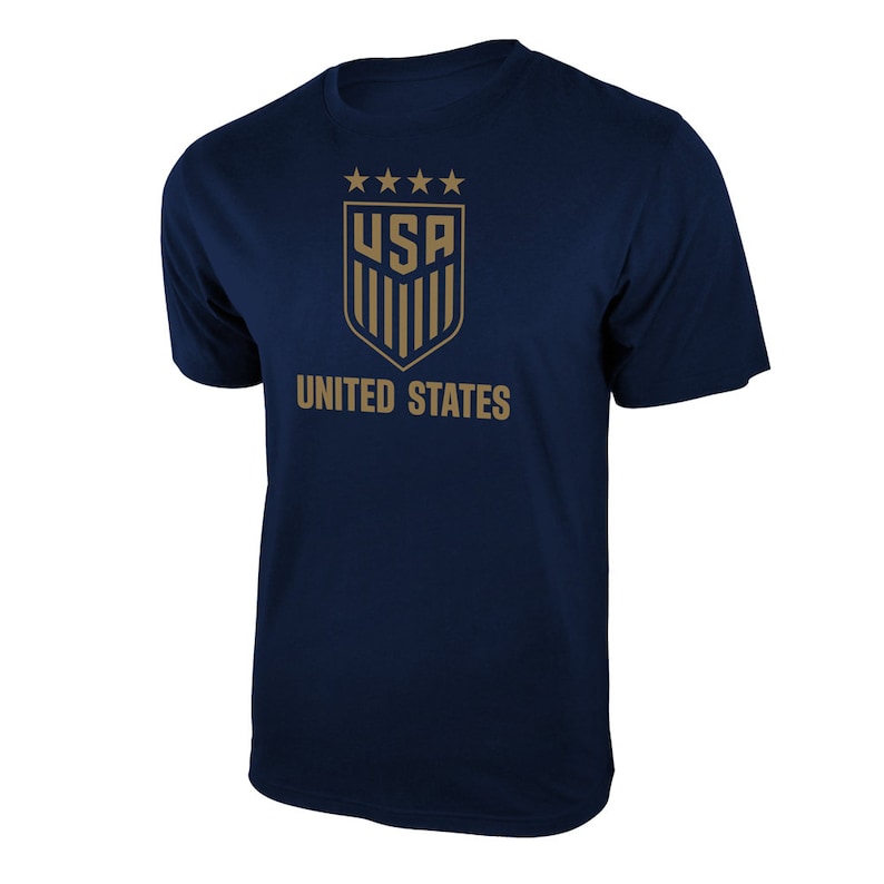 K&ouml;nnte beinhalten: Ein marineblaues T-Shirt mit einer goldenen Grafik des Logos der US-amerikanischen Fu&szlig;ballnationalmannschaft. Das Logo zeigt f&uuml;nf Sterne &uuml;ber den Buchstaben "USA" und einen Schild mit vertikalen Streifen. Die Worte "UNITED STATES" sind unter dem Logo gedruckt.