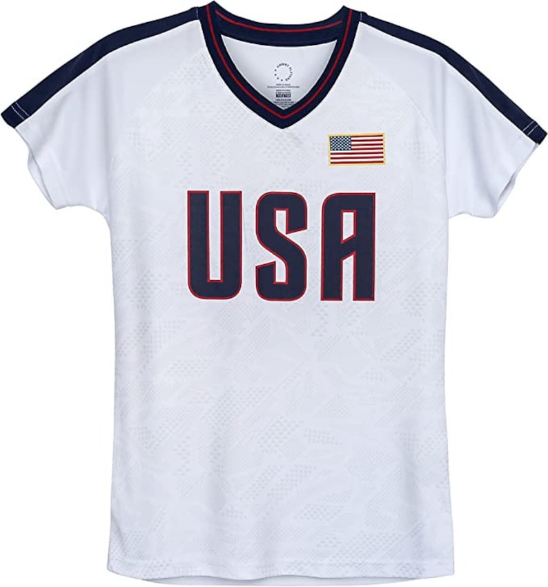 K&ouml;nnte beinhalten: Wei&szlig;es, kurz&auml;rmeliges Trikot mit dunkelblauem und rotem Besatz. Das Trikot hat einen V-Ausschnitt und tr&auml;gt den Schriftzug "USA" in Dunkelblau mit roter Umrandung. Auf der rechten Brustseite befindet sich eine kleine amerikanische Flagge.
